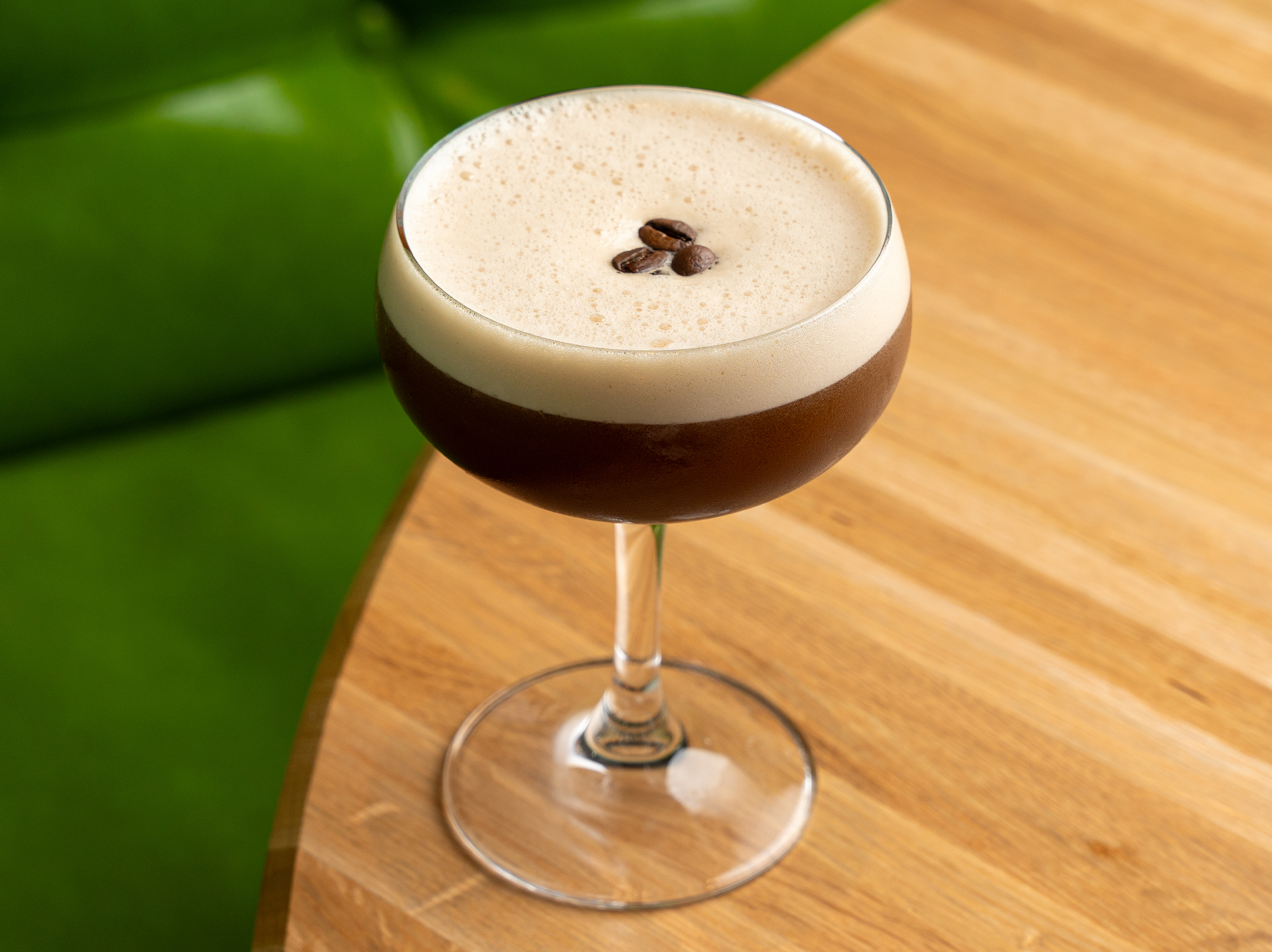 Espresso Martini from True Food Kitchen, featuring a creamy espresso cocktail in a martini glass, topped with frothy foam and three coffee beans, placed on a wooden table with a green cushioned background.