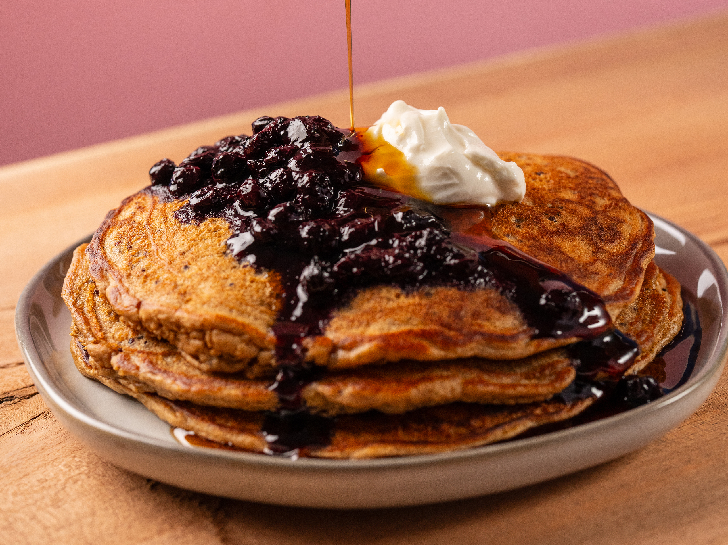 A stack of True Food Kitchen's golden-brown pancakes topped with a dollop of whipped cream and blueberry compote, with syrup being drizzled over them, set on a wooden table with a pink background.