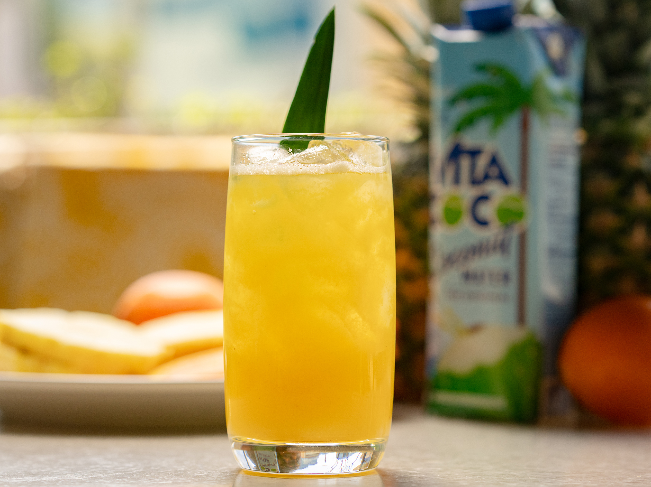 A tall glass of refreshing pineapple-coconut drink from True Food Kitchen, garnished with a pineapple leaf, with a background featuring a Vita Coco coconut water carton, a whole pineapple, and some oranges.