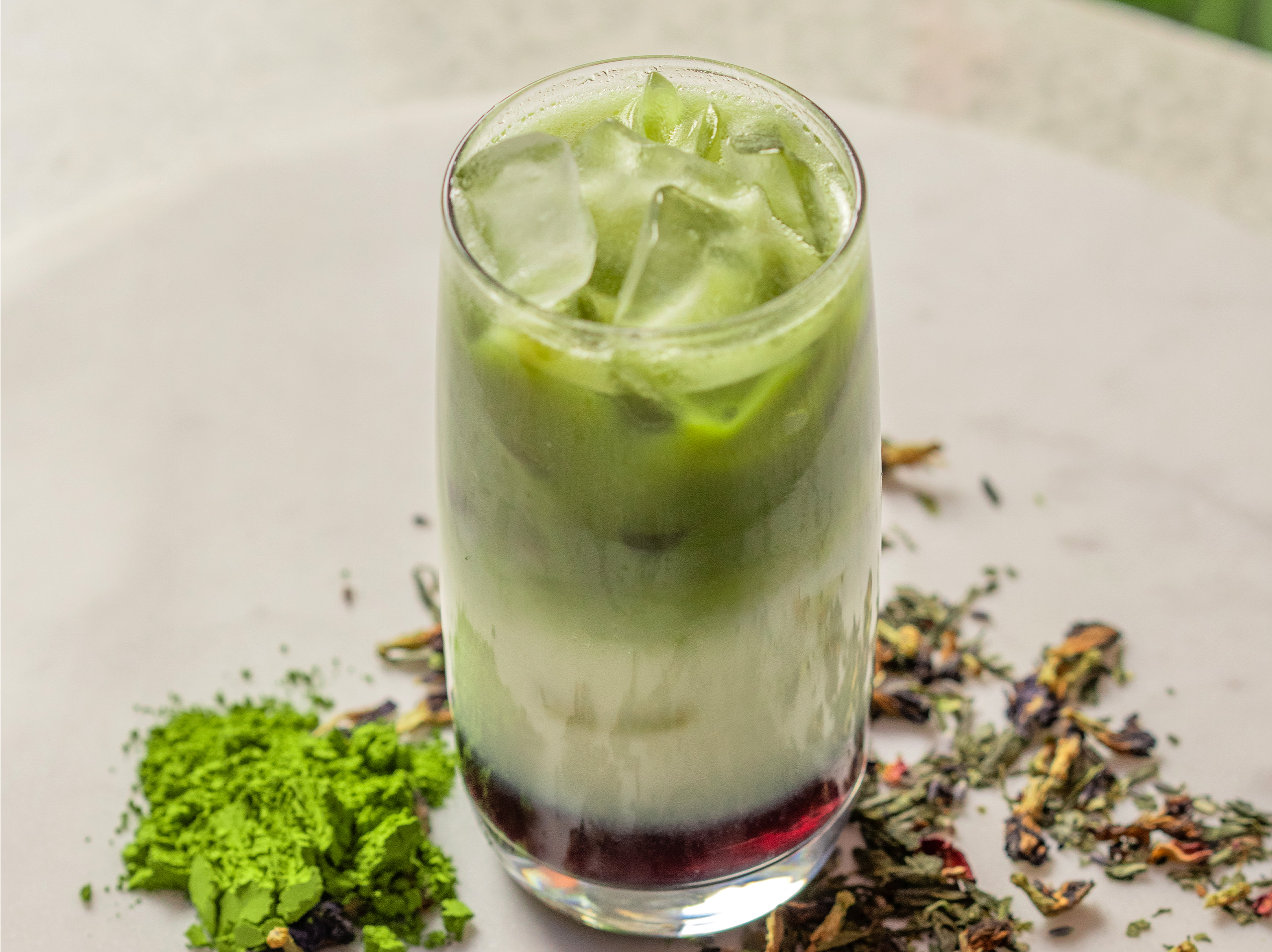 A refreshing iced matcha latte from True Food Kitchen, served in a tall glass with layers of vibrant green matcha, creamy milk, and a hint of berry syrup at the bottom, garnished with ice cubes and surrounded by loose leaf tea and matcha powder on a light surface.