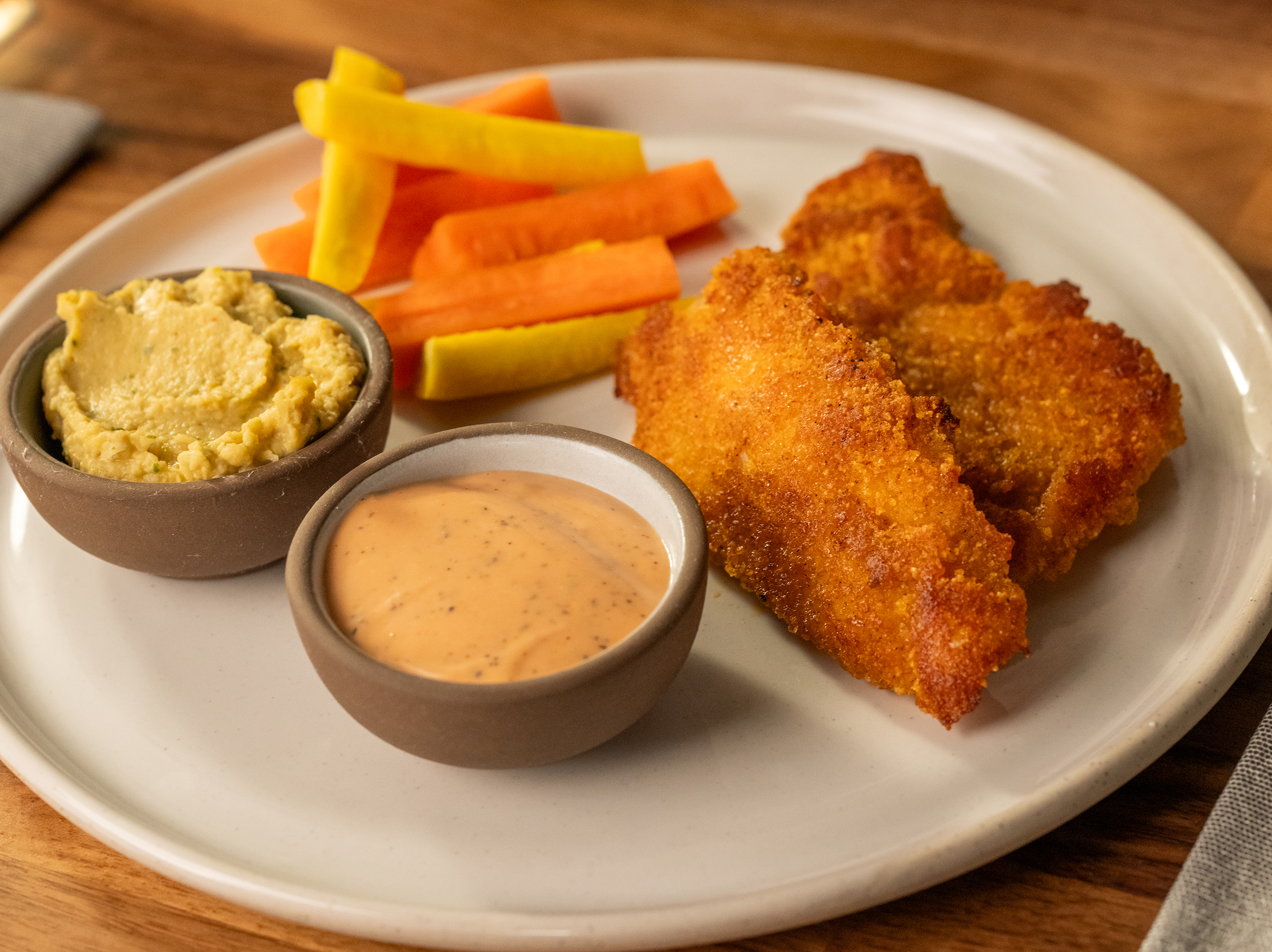 True Food Kitchen's kids menu dish featuring crispy chicken tenders, carrot and yellow vegetable sticks, with hummus and a creamy dipping sauce, served on a white plate.