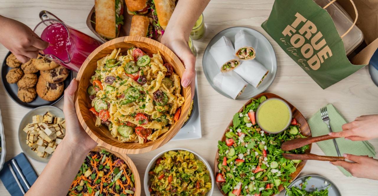 A person carrying a True Food Kitchen catering bag filled with fresh salads and side dishes, showcasing the convenience and variety of the catering options available.