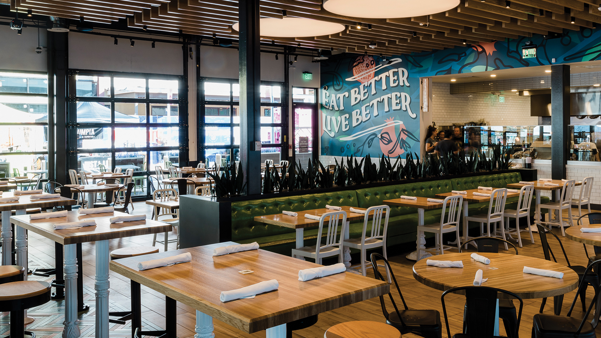 Interior view of a True Food Kitchen restaurant, featuring wooden tables neatly set with rolled napkins, metal chairs, and a green cushioned bench. The restaurant is bright and spacious with large windows, modern decor, and a vibrant mural on the wall that reads 'Eat Better, Live Better.