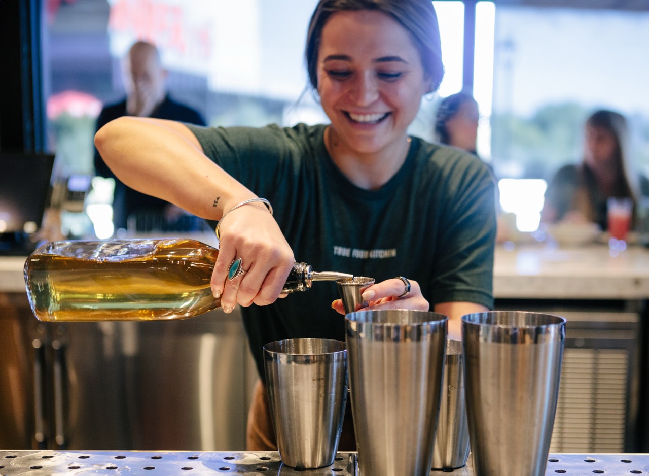 A True Food Kitchen staff member wearing a shirt that says 'Don't Kale My Vibe,' standing behind the bar, with a welcoming and energetic atmosphere in the background.